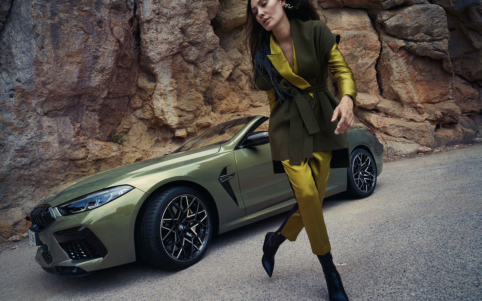 Atletiška priekinė dalis su M priekinio bamperio apdaila ir M serijos dvispalviai lengvojo lydinio dvispalviai ratlankiai 813 M BMW M8 Competition kabrioletas F91 LCI Facelift 2022 BMW Individual metalizuotos Brass kėbulo spalvos, stovinčio akmenuotoje vietovėje automobilio trijų ketvirčių vaizdas iš priekio su moterimi šalia