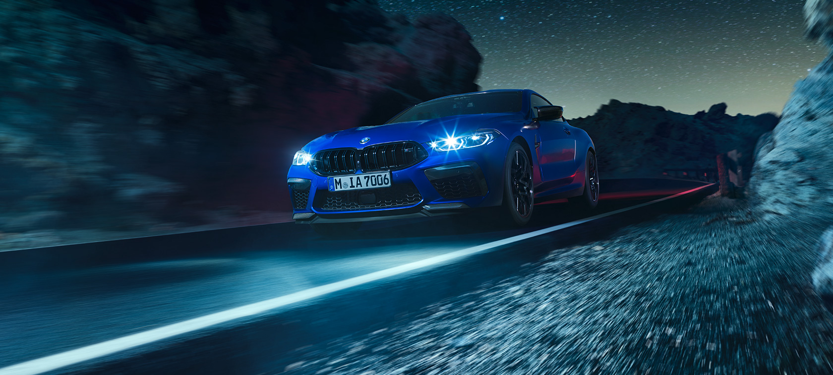BMW M8 Competition kupė, trijų ketvirčių vaizdas iš priekio BMW M8 Competition kupė Marina Bay Blue mėlyno metaliko spalvos, trijų ketvirčių naktį važiuojančio automobilio vaizdas iš priekio.