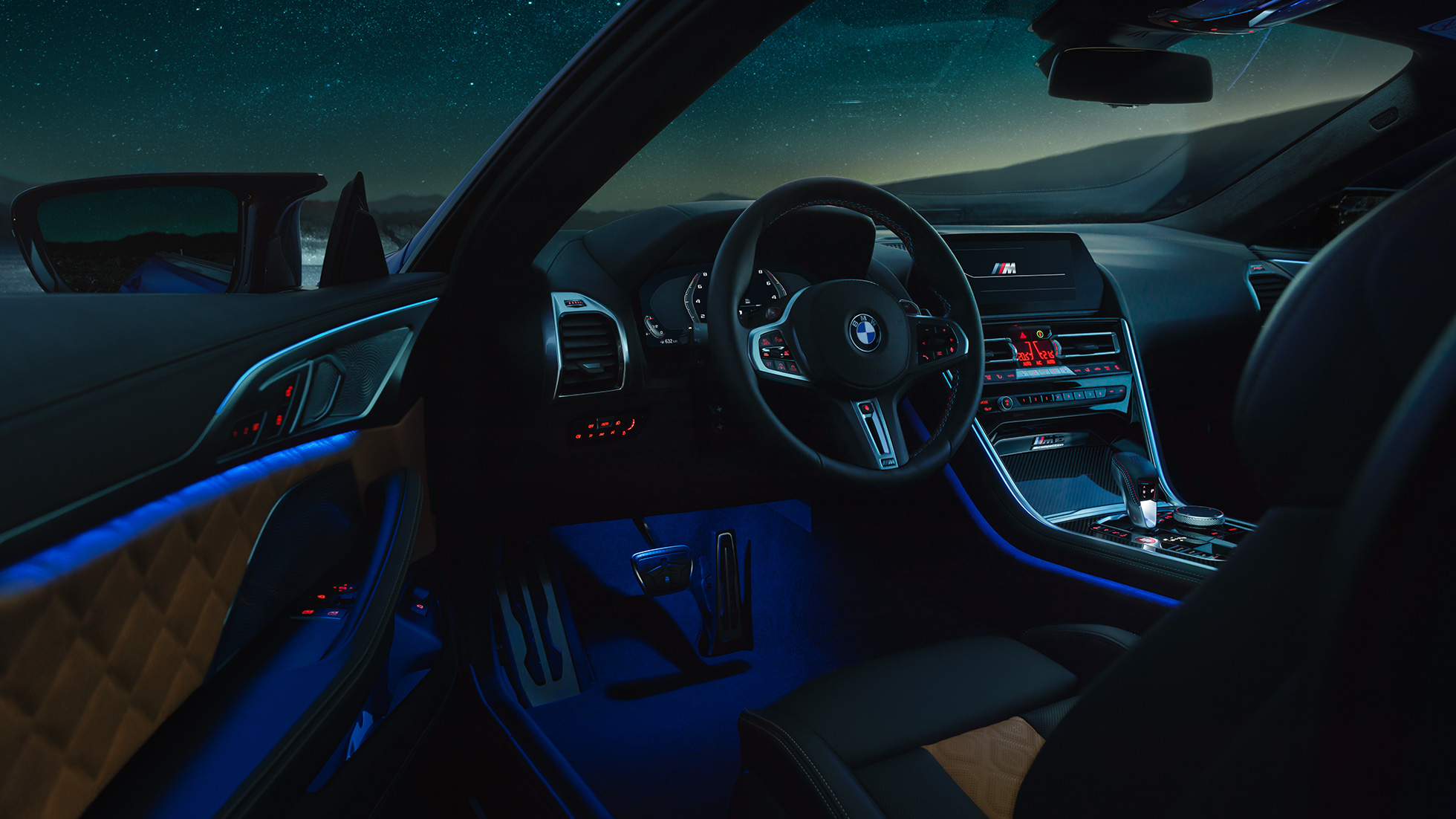 BMW M8 Competition kupė, interjeras BMW M8 Competition kupė, interjero su M odiniu vairu ir mėlynu aplinkos apšvietimu bei atviromis vairuotojo durelėmis vaizdas naktį