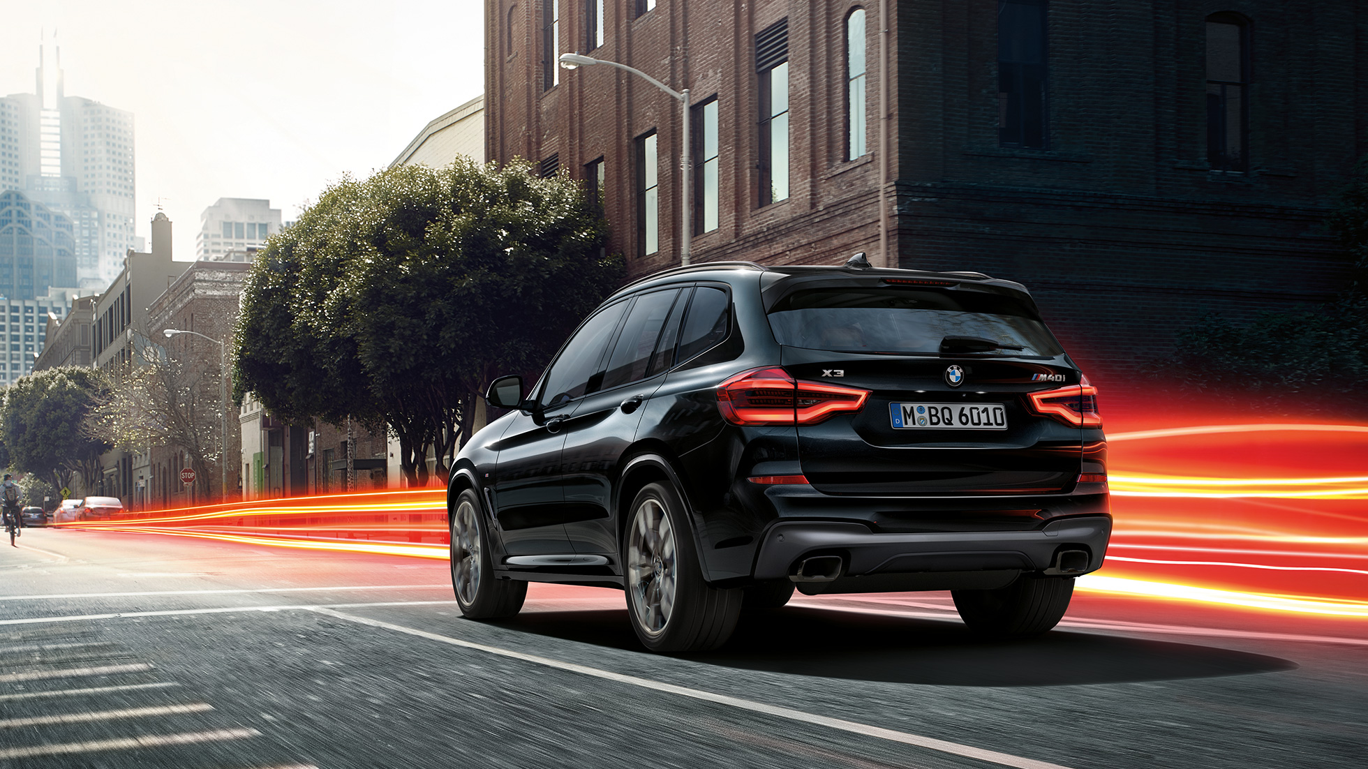 BMW X3 M40i, trijų ketvirčių vaizdas iš galo. BMW X3 M40i „Black Sapphire“ juodo metaliko spalvos, važiuojantis keliu, eksterjeras, trijų ketvirčių vaizdas iš galo apšviesto miesto fone.