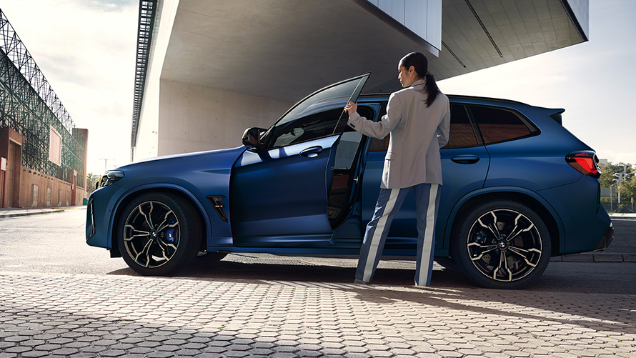 BMW X3 M Competition (F97) LCI Facelift 2021, automobilio atviromis durelėmis vaizdas iš šono su moterimi šalia BMW X3 M Competition (F97) LCI Facelift 2021 metalizuotos mėlynos Frozen Marina Bay Blue kėbulo spalvos, automobilio atviromis durelėmis vaizdas iš šono su moterimi šalia