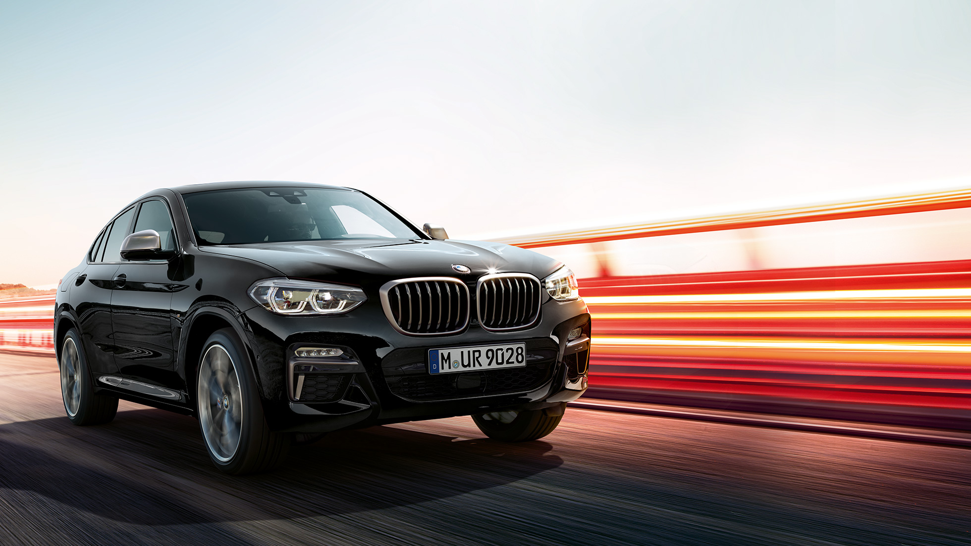 BMW X4 M40i, trijų ketvirčių vaizdas iš galo. BMW X4 M40i „Black Sapphire“ juodo metaliko spalvos, važiuojantis keliu, eksterjeras, trijų ketvirčių apšviestas vaizdas iš galo.