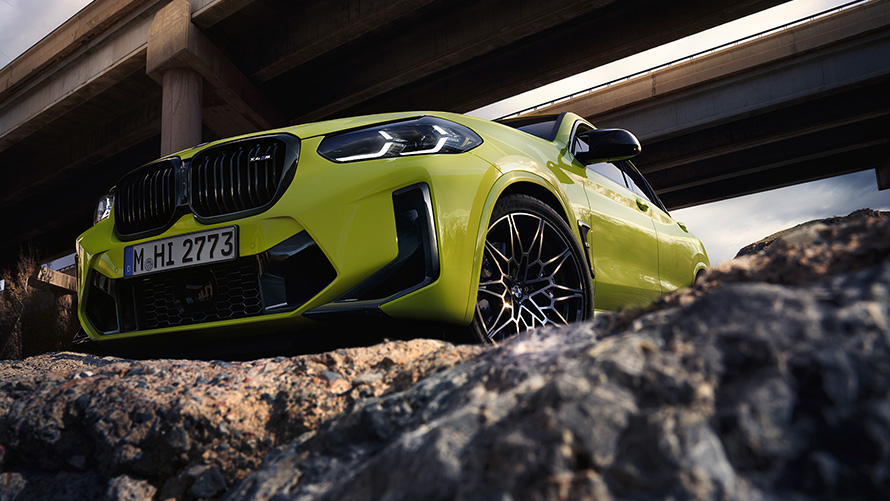BMW X4 M Competition (F98) LCI 2021 trijų ketvirčių vaizdas iš priekio BMW X4 M Competition (F98) LCI Facelift 2021 geltonos Sao Paulo Yellow spalvos po tiltu važiuojančio automobilio trijų ketvirčių vaizdas iš priekio