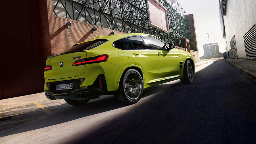 BMW X4 M Competition (F98) LCI 2021 trijų ketvirčių vaizdas iš priekio BMW X4 M Competition (F98) LCI Facelift 2021 geltonos Sao Paulo Yellow spalvos trijų ketvirčių vaizdas iš priekio gamyklų fone