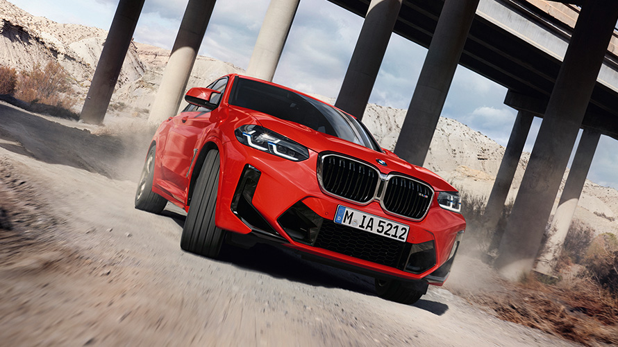 BMW X4 M (F98) LCI Facelift 2021 raudonos metalizuotos Toronto Red kėbulo spalvos trijų ketvirčių vaizdas iš priekio tilto kolonų fone