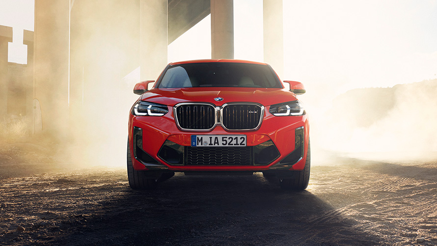 BMW X4 M (F98) LCI Facelift 2021 raudonos metalizuotos Toronto Red spalvos trijų ketvirčių vaizdas iš priekio tilto kolonų fone