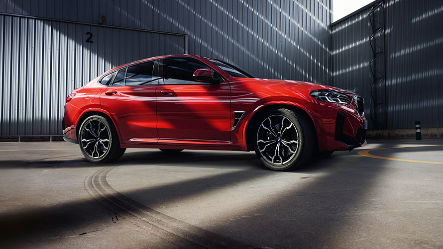 BMW X4 M (F98) LCI Facelift 2021 raudonos metalizuotos Toronto Red kėbulo spalvos trijų ketvirčių vaizdas iš šono gamyklos pastato fone