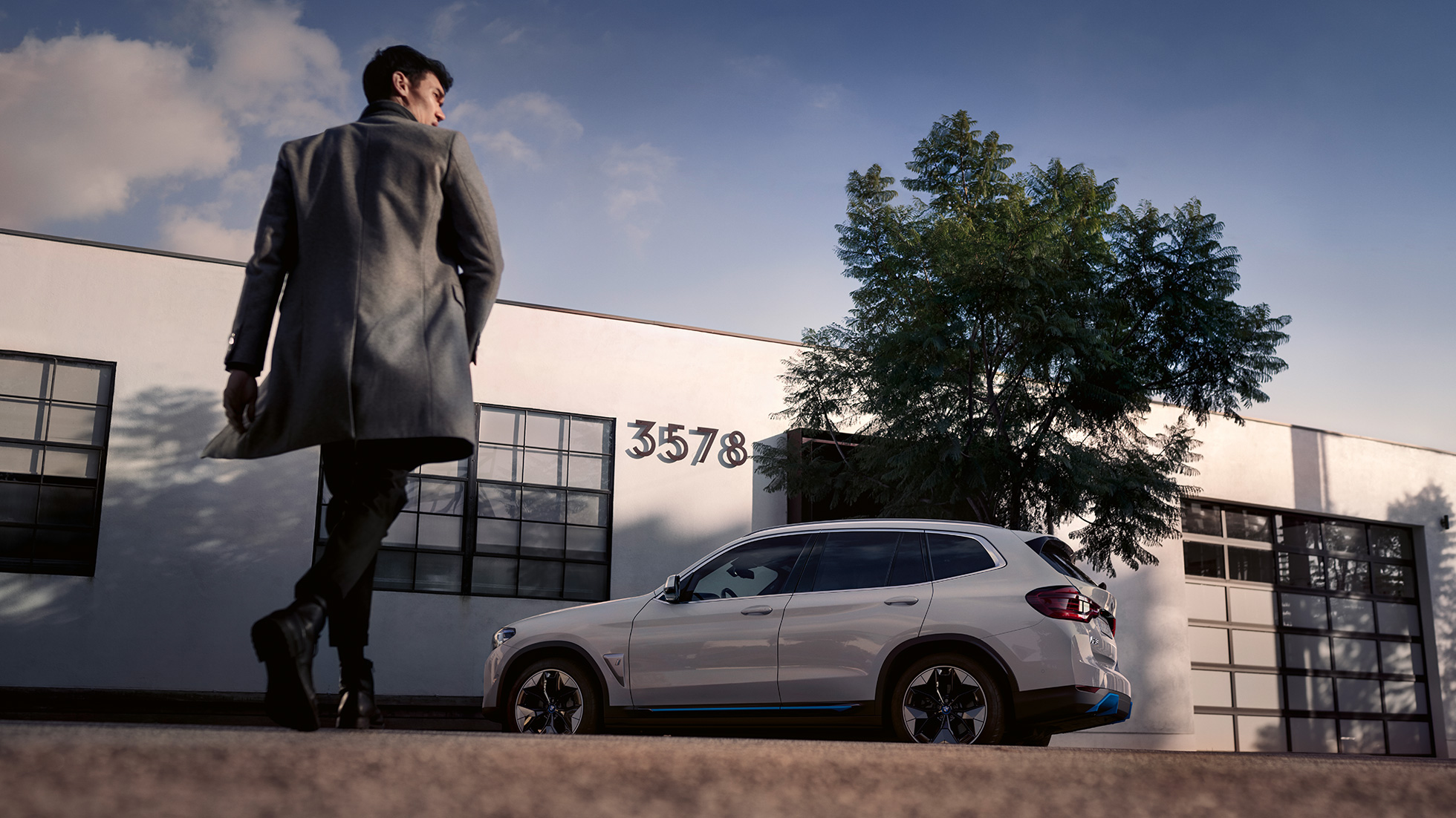 BMW iX3 G08, vaizdas iš šono prie pastato su vyru BMW iX3 G08 2020 balto Mineral White metaliko spalvos, vaizdas iš šono prie pastato su vyru