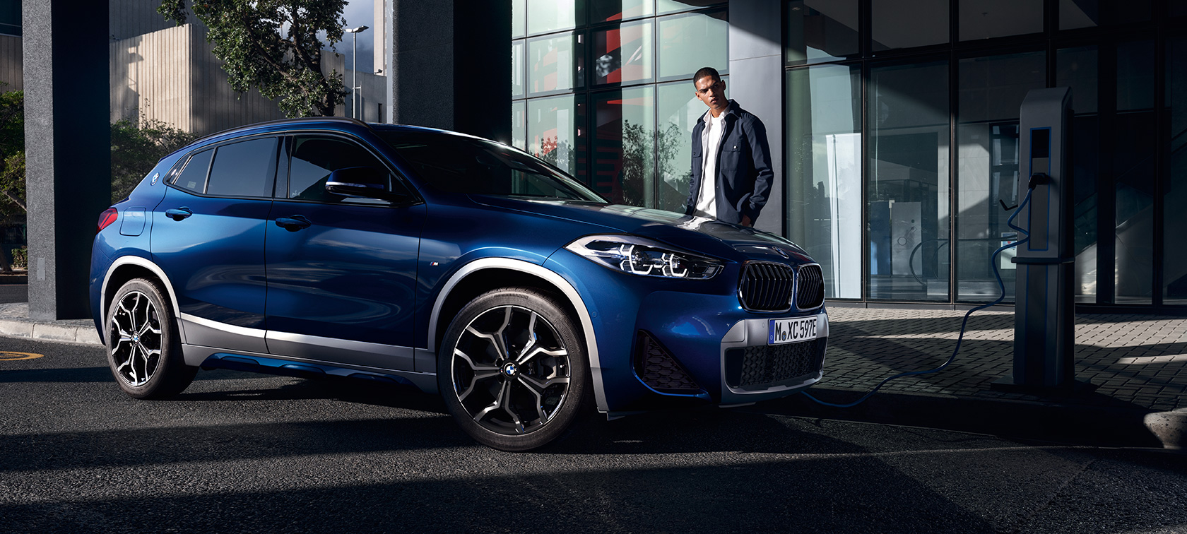 Įkraunamas hibridinis BMW X2 xDrive25e F39, automobilio trijų ketvirčių vaizdas iš šono įkrovimo stotelėje Įkraunamas hibridinis BMW X2 xDrive25e F39, automobilio trijų ketvirčių vaizdas iš šono įkrovimo stotelėje su modeliu