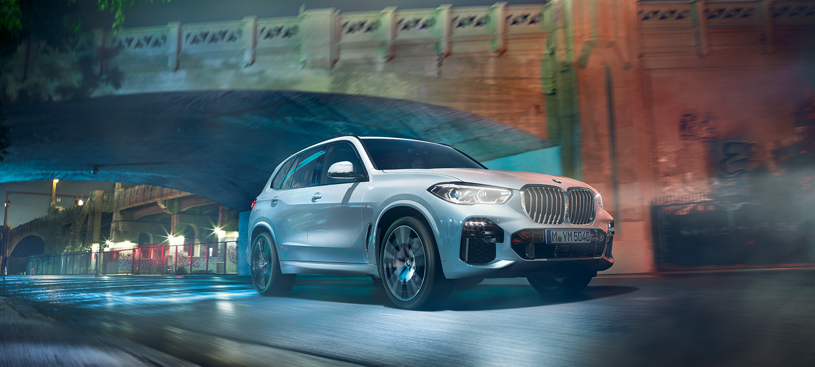 BMW X5 (G05) 2018 baltos metalizuotos Mineral White spalvos, nakties fone važiuojančio automobilio trijų ketvirčių vaizdas iš priekio
