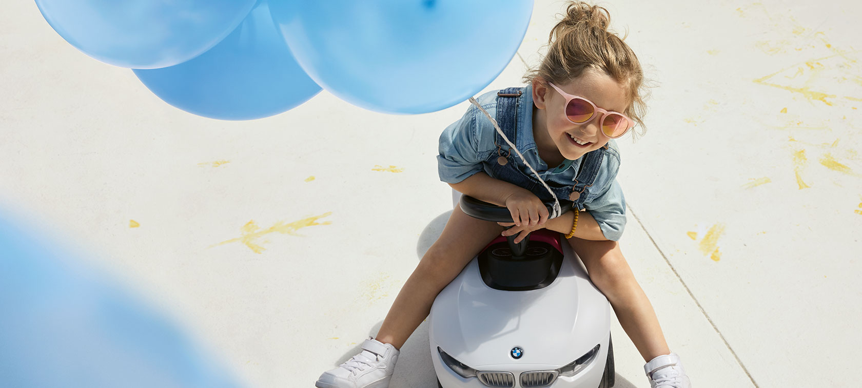 BMW vaikams Laiminga mergaitė rožiniais akiniais nuo saulės laiko mėlyną balioną ir sėdi BMW Baby Racer kūdikių mašinėlėje.