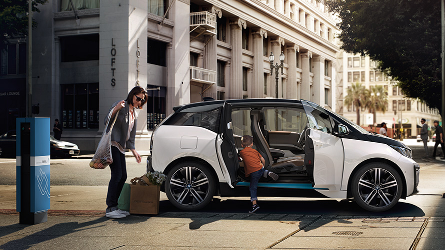 BMW i3 moteris ir vaikas miesto fone BMW nuvažiuojamas atstumas BMW i3 miesto fone su moterimi ir vaiku