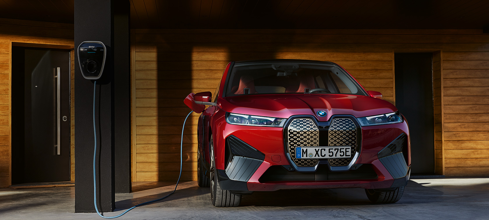Namuose sieninėje stotelėje įkraunamo BMW iX vaizdas iš priekio Namuose įrengtoje Wallbox įkrovimo stotelėje kraunamas BMW iX automobilis, stovintis priešais medinę garažo sieną