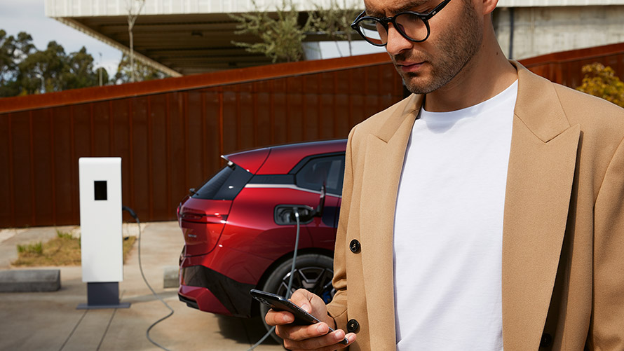 Moteris su išmaniuoju telefonu šalia BMW iX3 įkrovimo stotelėje Moteris su išmaniuoju telefonu šalia BMW iX3 įkrovimo stotelėje