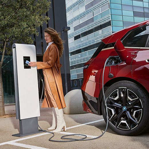 BMW iX i20 įkrovimo stotelėje su moterimi ir BMW Charging įkrovimo kortele