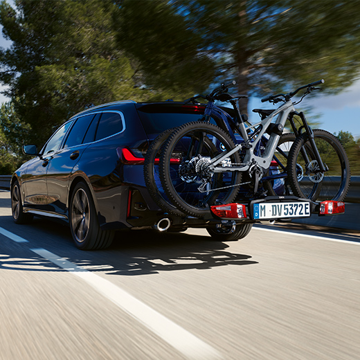 BMW 3 serijos Touring G21 PHEV 3/4 vaizdas iš galo su šalia važiuojančiais dviračiais. 