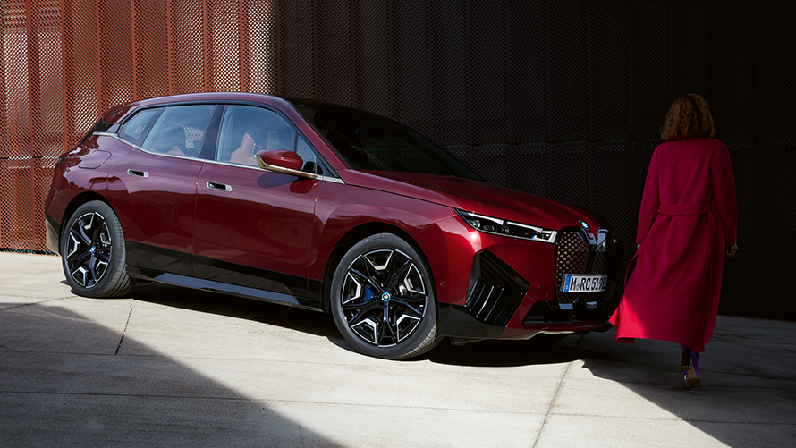 Moteris BMW iX i20 2021 priešakyje, trijų ketvirčių vaizdas iš priekio Moteris vilkinti raudonu paltu BMW iX i20 2021 priešakyje, trijų ketvirčių vaizdas iš priekio