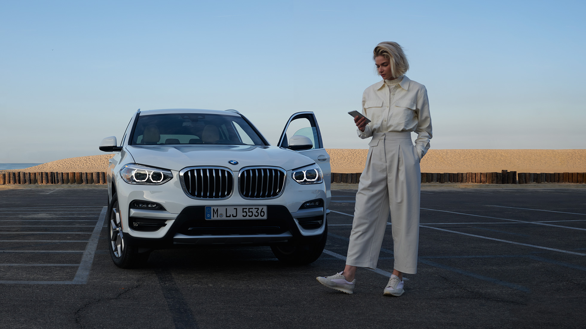 Moteris naudojasi My BMW programėle savo išmaniajame telefone My BMW programėle iš BMW ConnectedDrive savo išmaniajame telefone besinaudojanti moteris, stovinti baltos spalvos BMW X5 automobilio priešakyje