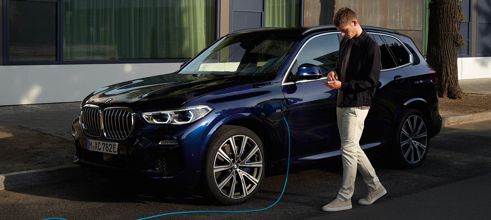 Vyras naudojasi My BMW programėle savo išmaniajame telefone My BMW programėle iš BMW ConnectedDrive savo išmaniajame telefone besinaudojantis vyras, kraunantis savo įkraunamą BMW X5 hibridą