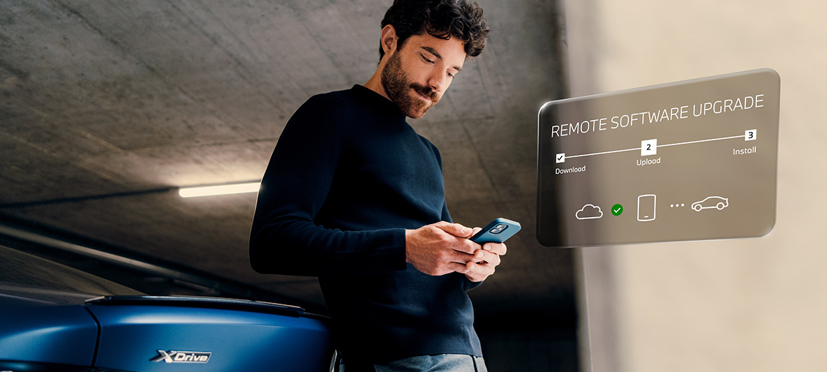 Į galinę BMW automobilio dalį atsirėmęs vyras, žvelgiantis į savo išmanųjį telefoną Nuotolinis programinės įrangos atnaujinimas, į galinę BMW automobilio dalį atsirėmęs vyras, žvelgiantis į savo išmanųjį telefoną