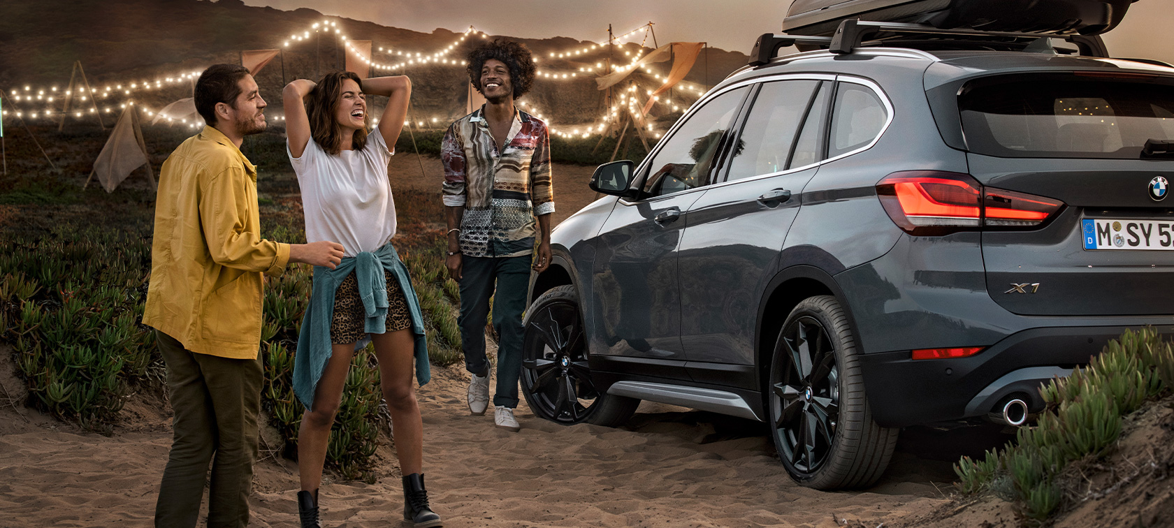 Laiminga trijulė BMW X1 automobilyje su BMW vasaros pasiūlymų įranga Laiminga trijulė BMW Mineral Grey spalvos BMW X1 automobilyje su BMW vasaros pasiūlymų įranga; vaizdas iš galo paplūdimyje, žvelgiant trijų ketvirčių kampu