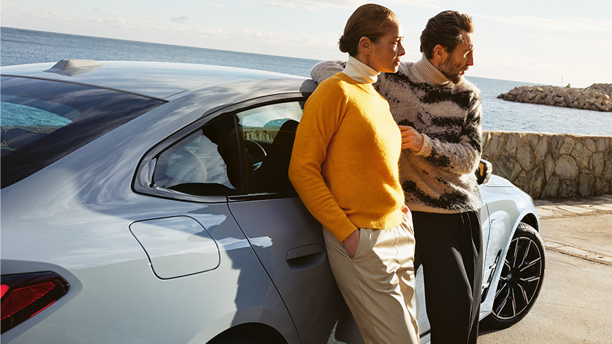 BMW Service Inclusive paslaugų teikimo trukmė naujiems automobiliams ir taikomi sutarčių galiojimo terminai BMW Service Inclusive paslaugos, priešais BMW i4 automobilį pakrantėje stovintys vyras ir moteris