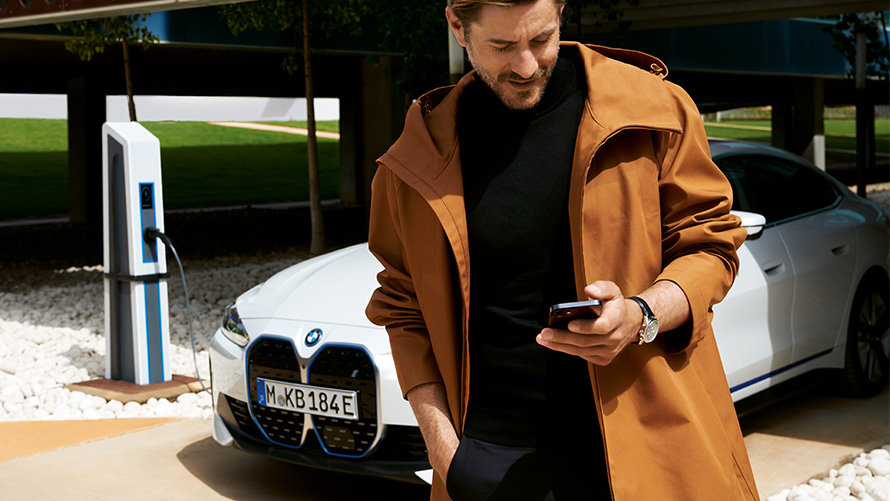 BMW Service Inclusive paslaugų teikimo trukmė ir elektriniams automobiliams taikomi sutarčių galiojimo terminai BMW Service Inclusive paslaugos, priešais BMW i4 automobilį įkrovimo stotelėje stovintis vyras, žvelgiantis į išmanųjį telefoną