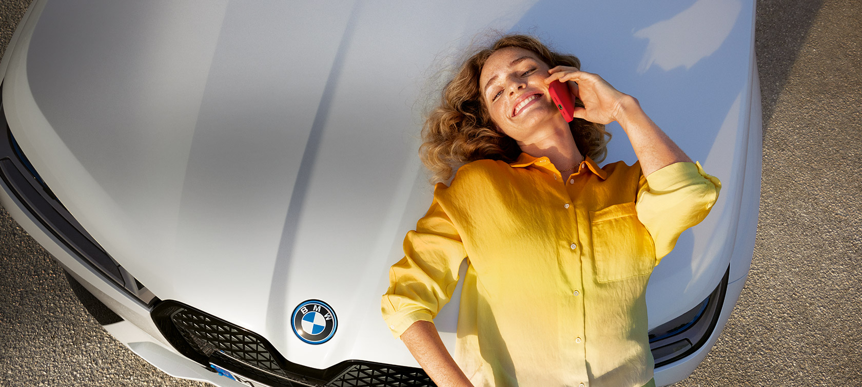 Relax We Care iniciatyvioji priežiūros paslauga Proactive Care, patikra ir remontas Proactive Care paslauga, automobilio patikra ir remontas, Relax We Care, moteris geltonais marškiniais ir išmaniuoju telefonu palinkusi prie balto BMW variklio dangčio