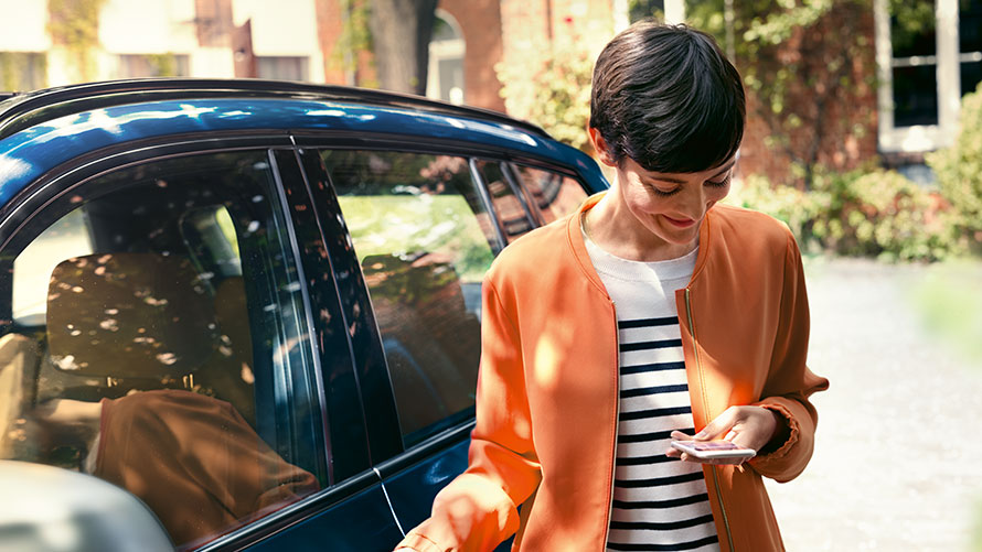 BMW telepaslaugos BMW telepaslaugos: moteris stovi priešais BMW ir naudojasi išmaniuoju telefonu