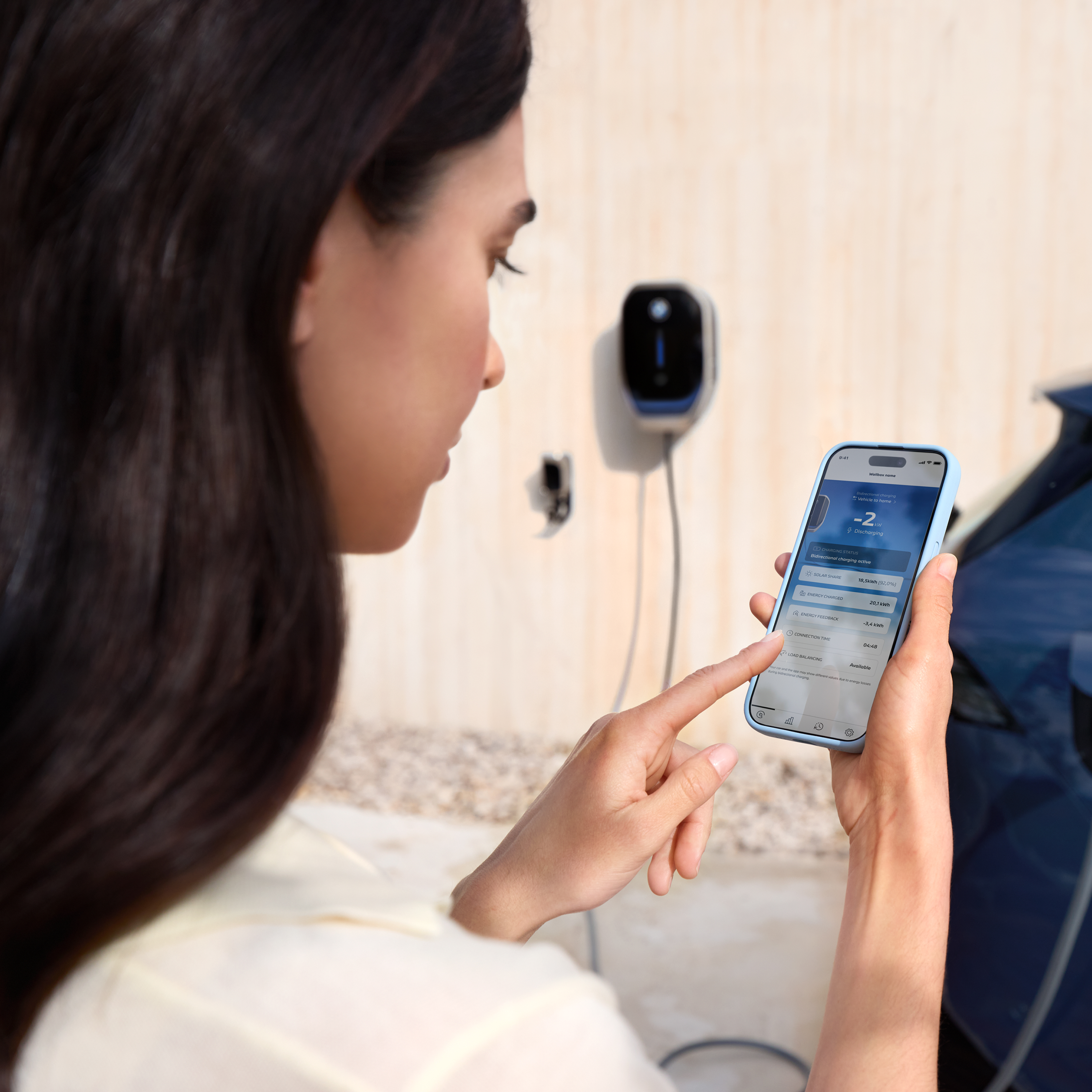 Moteris žiūri į savo išmanųjį telefoną Wallbox sieninės įkrovimo stotelės fone